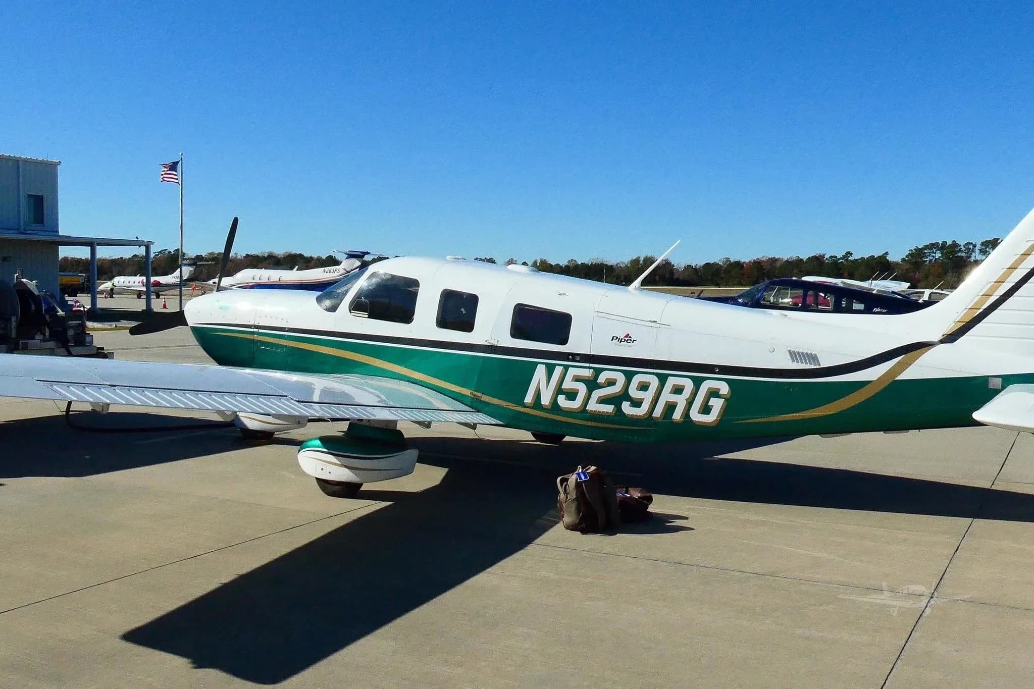 A side view of the white and green Piper 6XT (N529RG) parked on an airport tarmac with a building and other aircraft in the background.