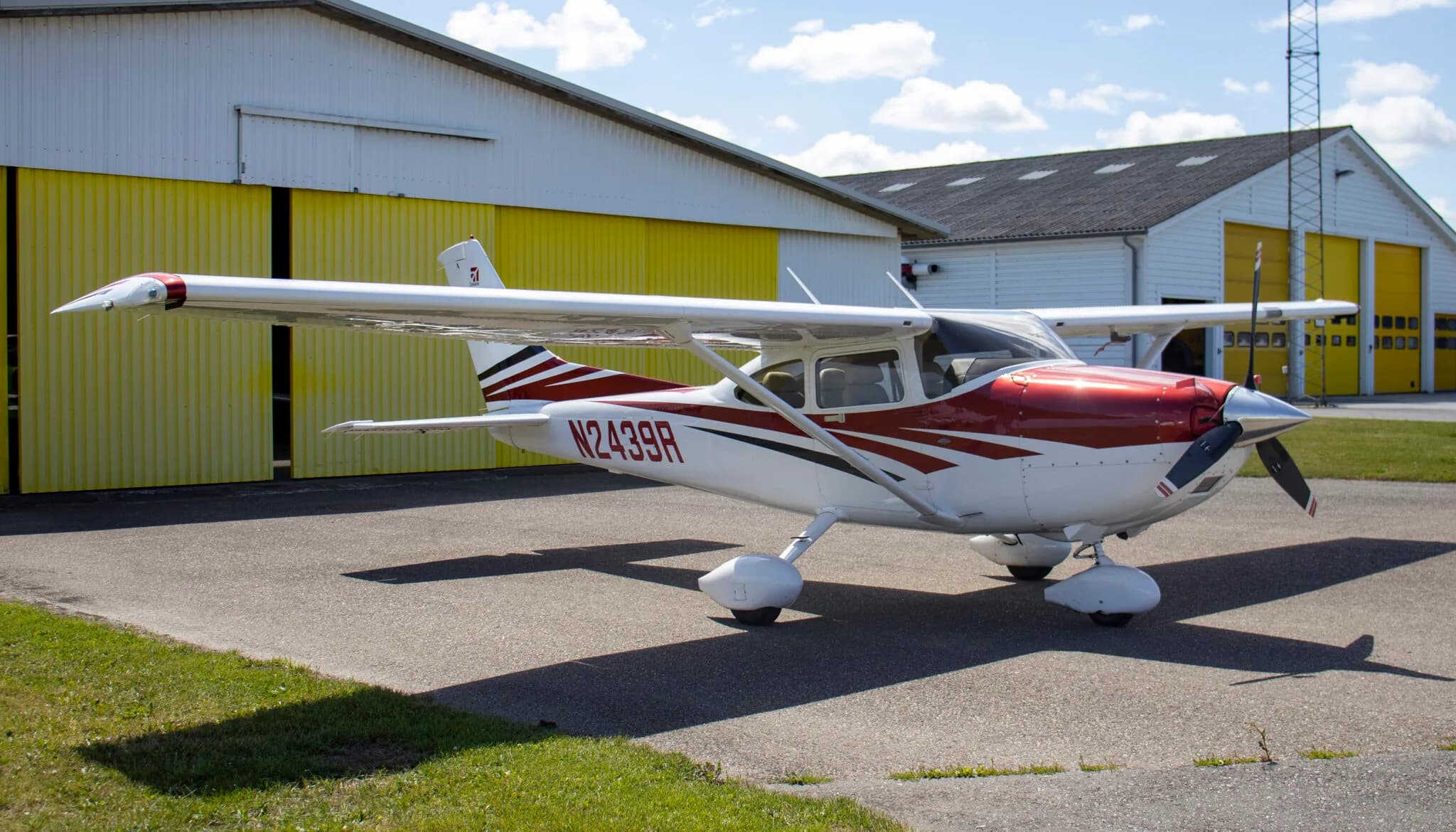 A white, red, and black Cessna 182 aircraft with registration N2439R parked on an airfield in front of hangars.