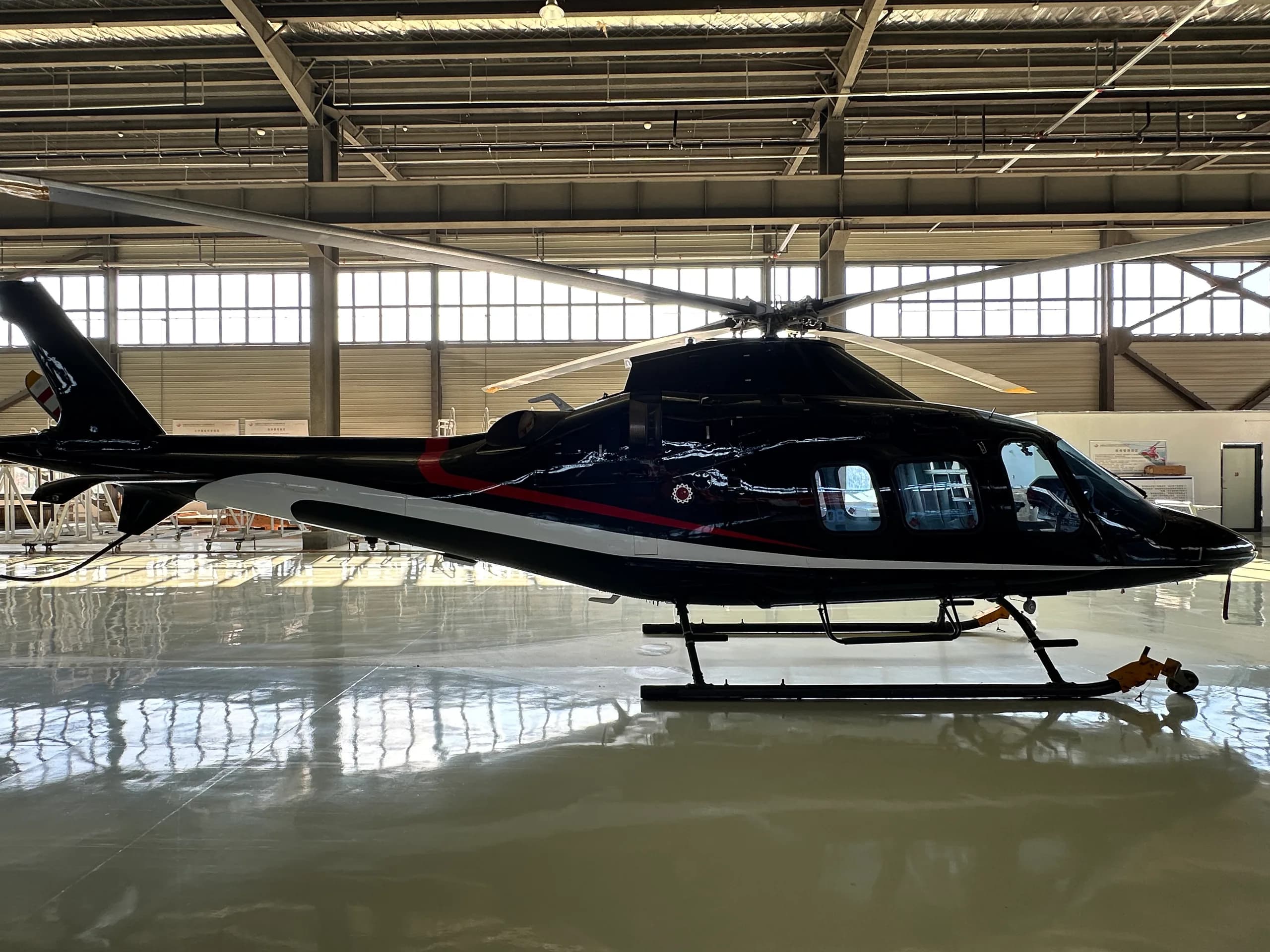 A black helicopter with white and red stripes is parked on a reflective floor in a hangar, viewed from the left side.