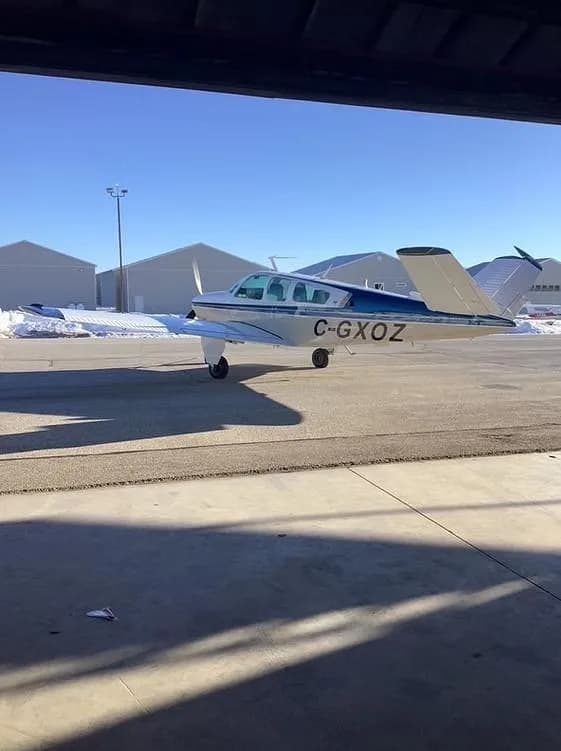 A beige and blue single-engine aircraft with registration C-GXOZ is parked on an airfield, viewed from inside a hangar.