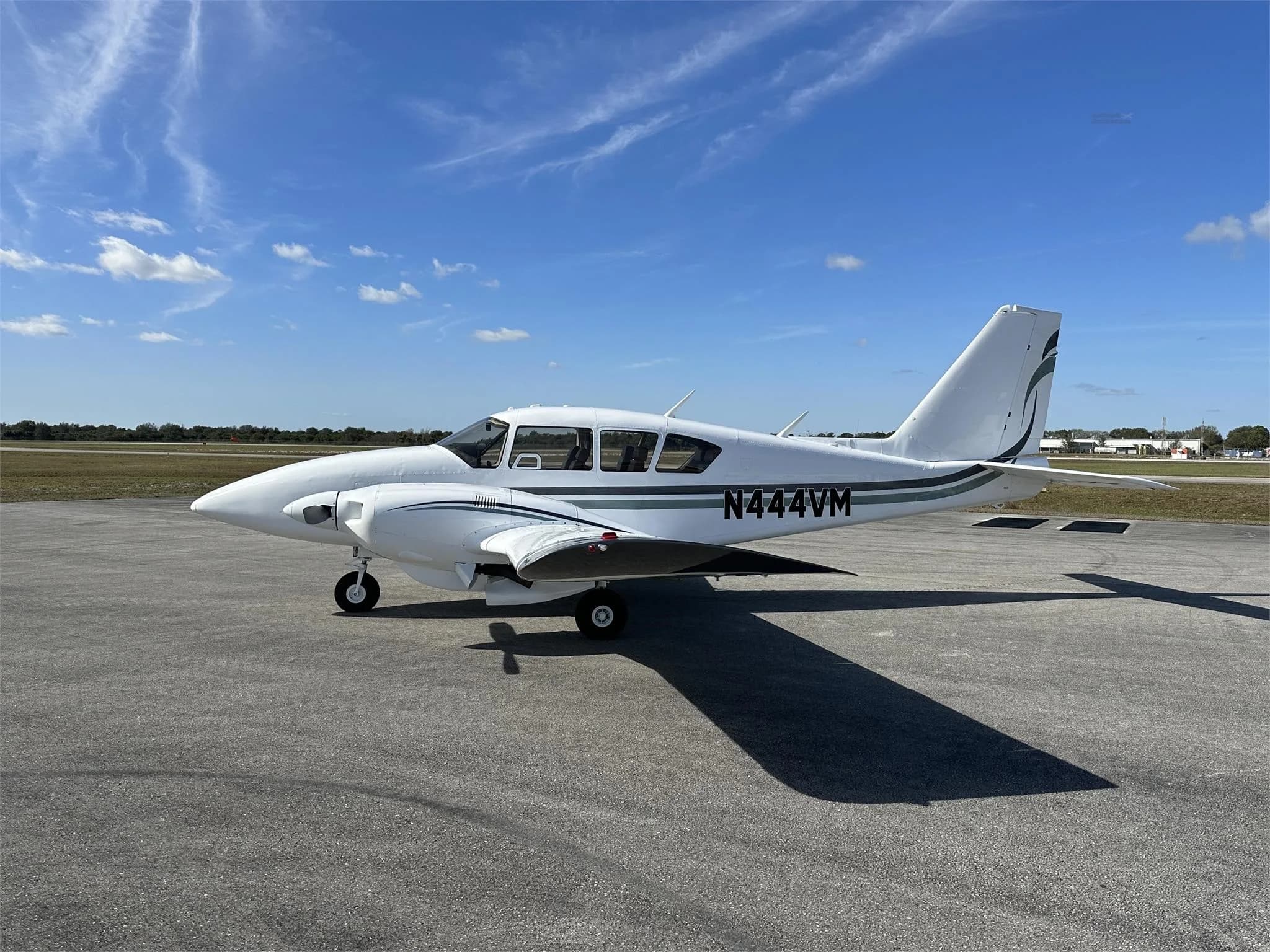 A side view of a white Piper aircraft with green and black stripes, registration N444VM, parked on an airfield.