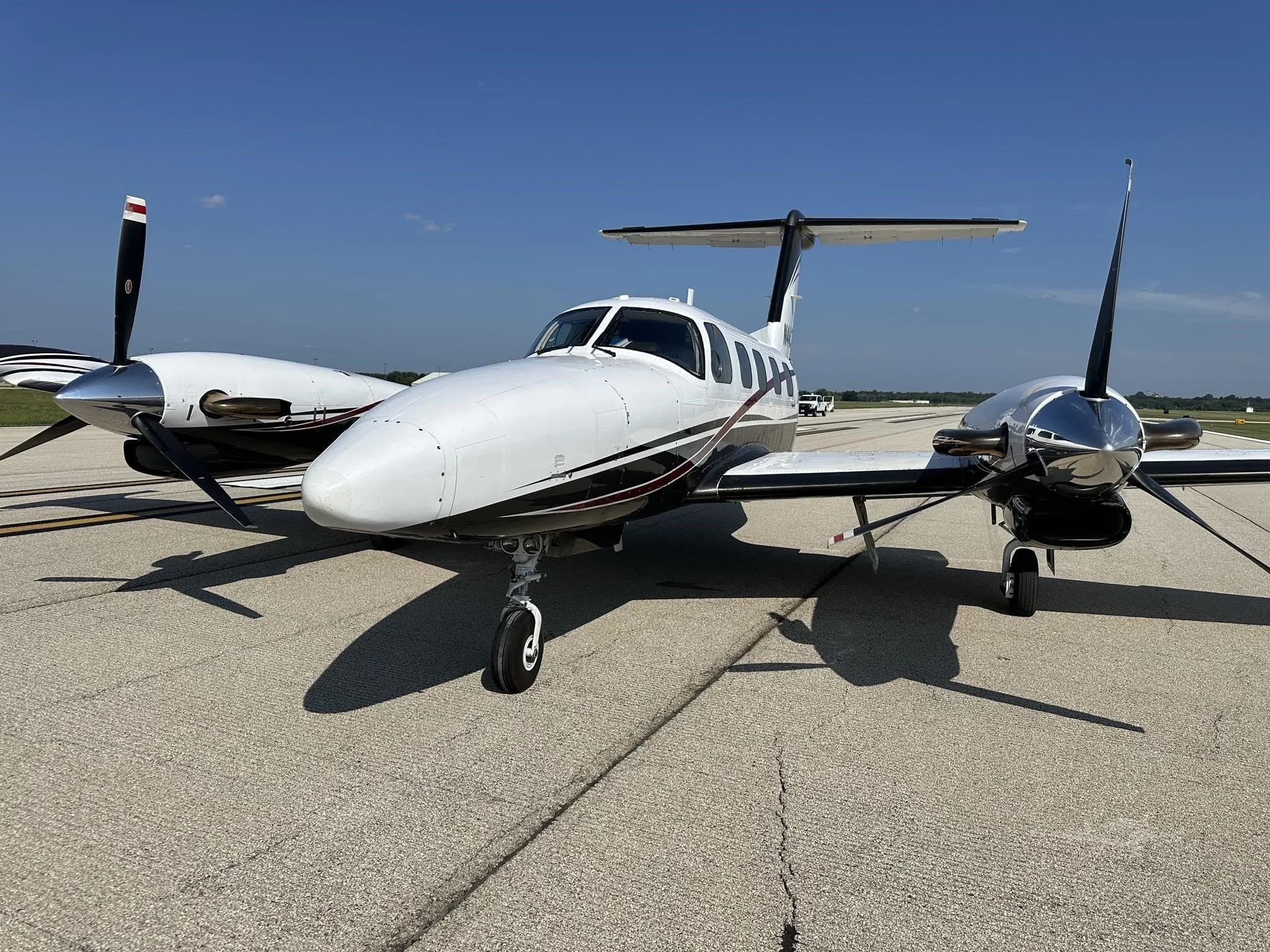 A head-on view of a white and dark grey twin-engine turboprop aircraft with red pinstripes, tail number N444CY, parked on an airport runway under a clear blue sky.