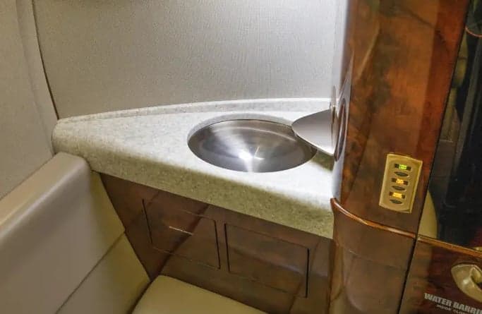 A close-up view of a small sink area with a light-colored countertop and dark wood cabinetry inside an aircraft.