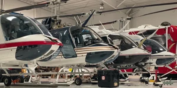 Multiple helicopters with various paint schemes are lined up inside a hangar, some appearing to be under maintenance.