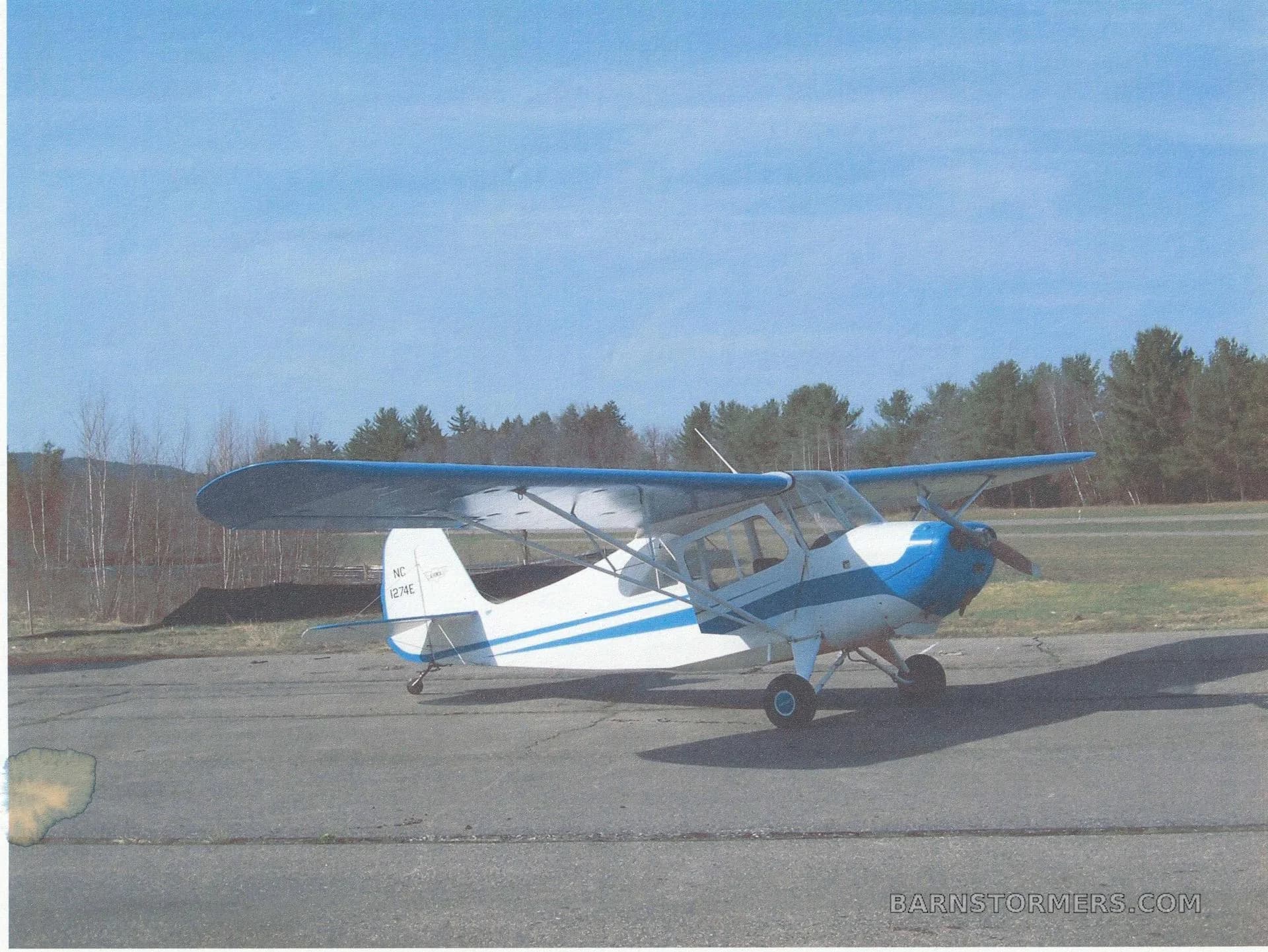 A white and blue single-engine high-wing aircraft, registration NC 1274E, parked on an asphalt ramp with trees in the background.