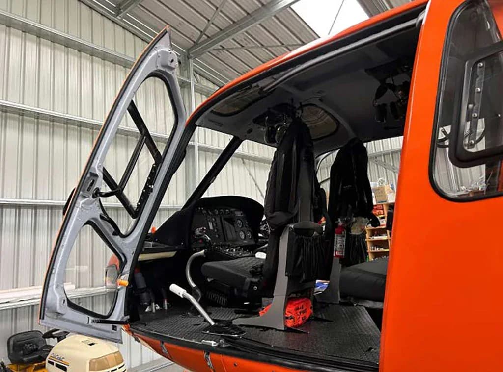 A view into the open front door of an orange helicopter, showing the pilot's seat, controls, and part of the instrument panel.