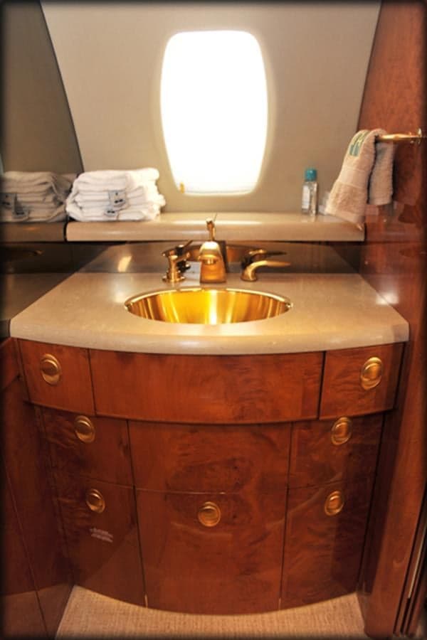 A private jet lavatory features a polished gold sink, a light-colored countertop, and rich wood cabinetry below a window.