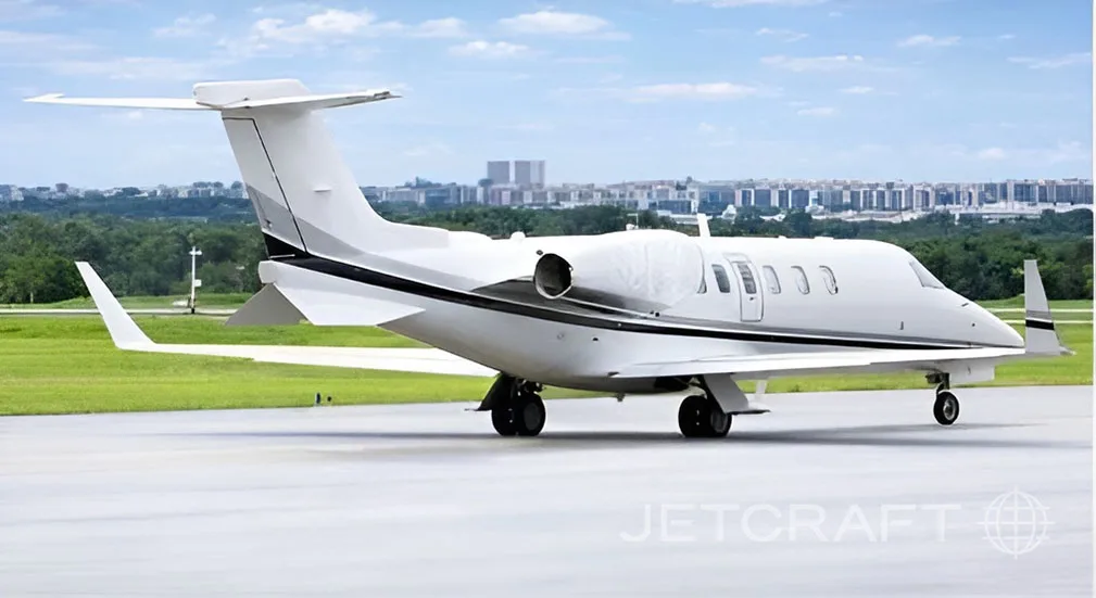 A white business jet with a black and grey stripe parked on the tarmac with a city skyline in the background.
