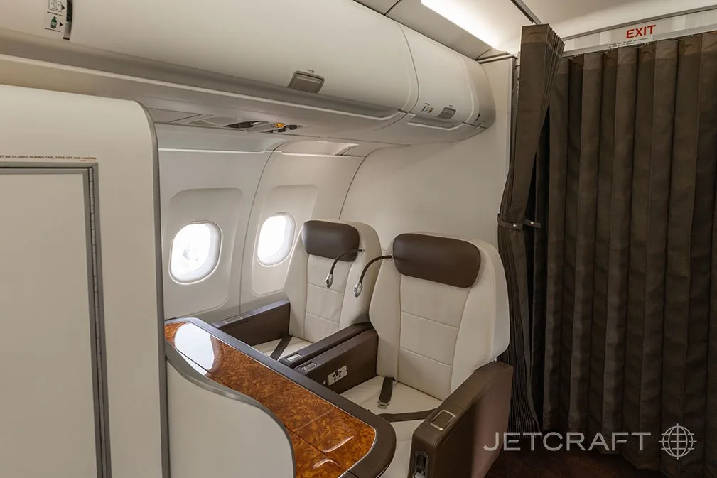 A private aircraft cabin section with two cream and brown leather seats next to windows, separated by a curtain.