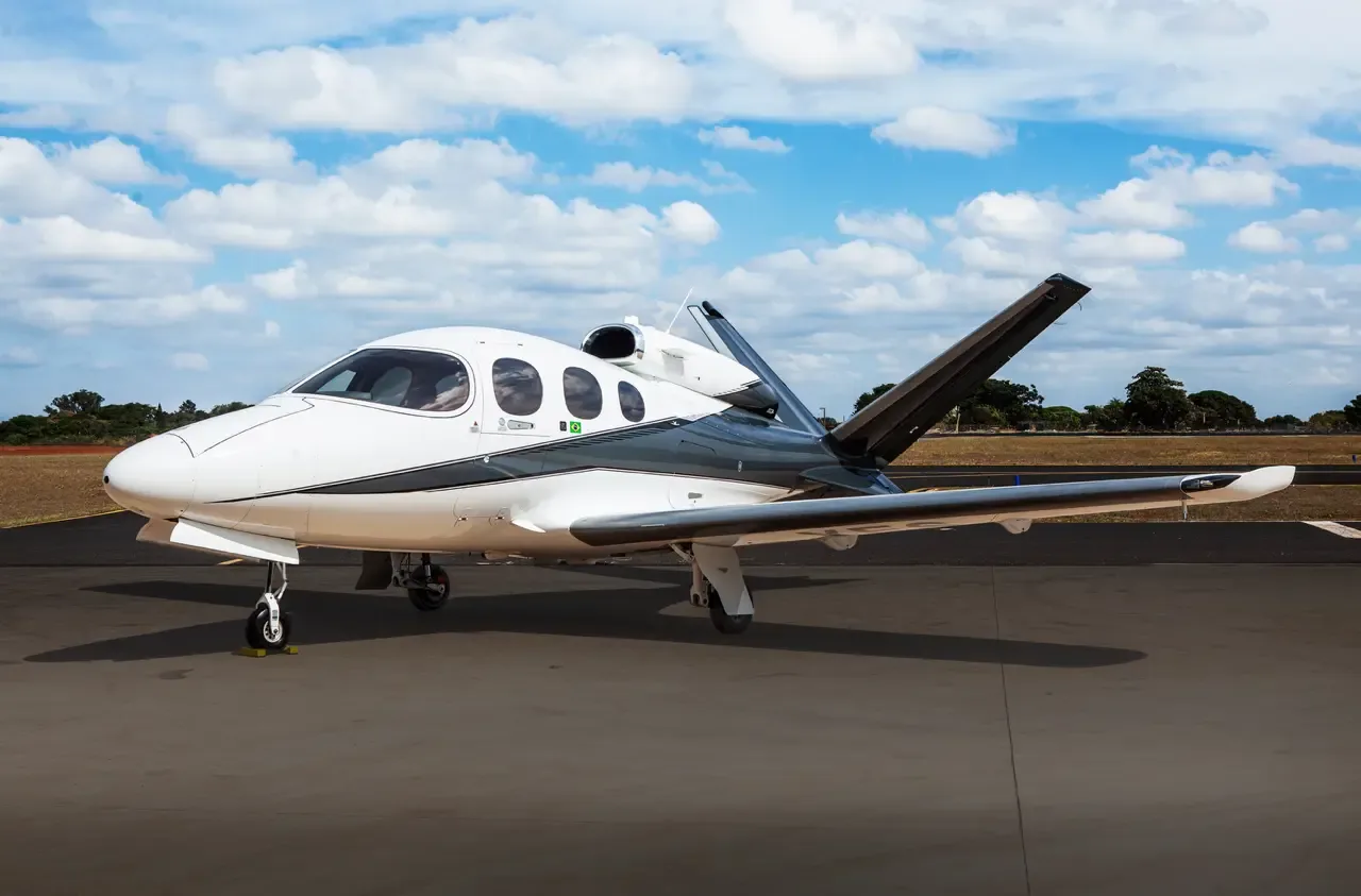 A white and dark grey Cirrus Vision Jet parked on an asphalt tarmac under a partly cloudy sky.