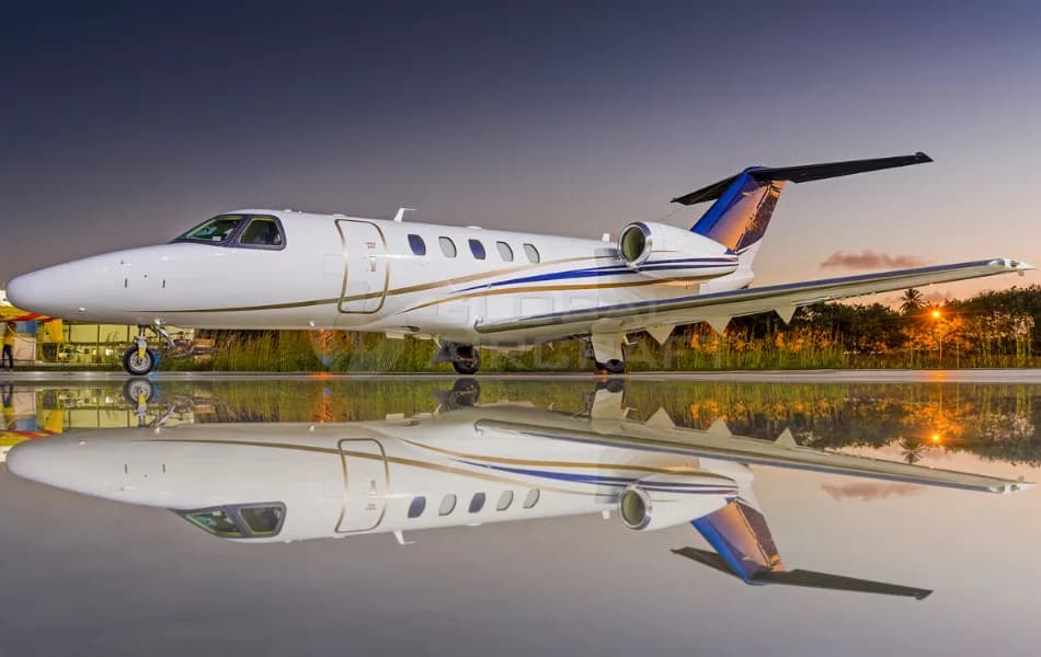A white business jet with gold and blue stripes and a colorful tail, parked on a reflective surface at dusk.