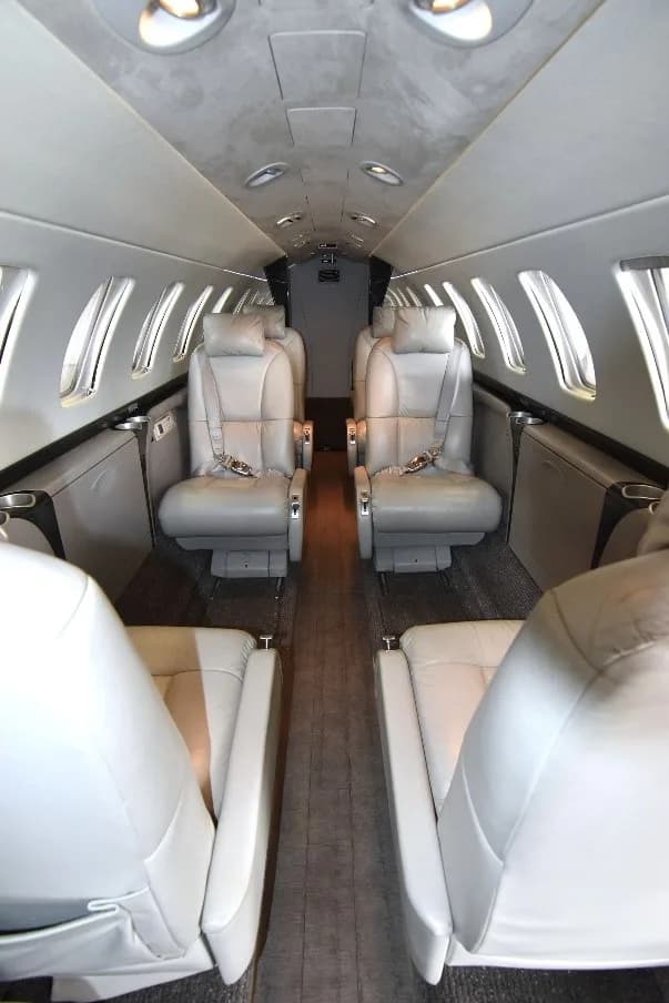 The interior cabin of a private jet, showing light gray leather seats and the aisle from a rear perspective.