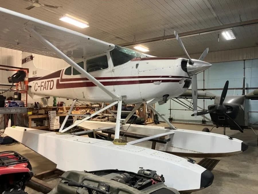 A white floatplane with maroon stripes, registration C-FATD, is parked inside a hangar with another aircraft in the background.