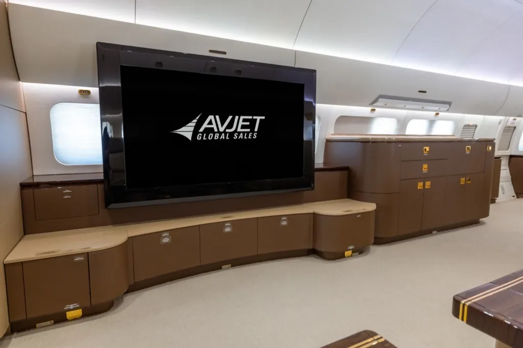 A large flat-screen television displaying the Avjet Global Sales logo, mounted above brown cabinetry in an aircraft cabin.