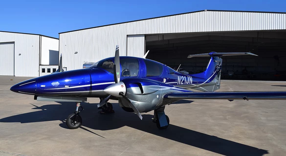 A Diamond DA62 aircraft with a blue and silver paint scheme is parked outside a hangar.