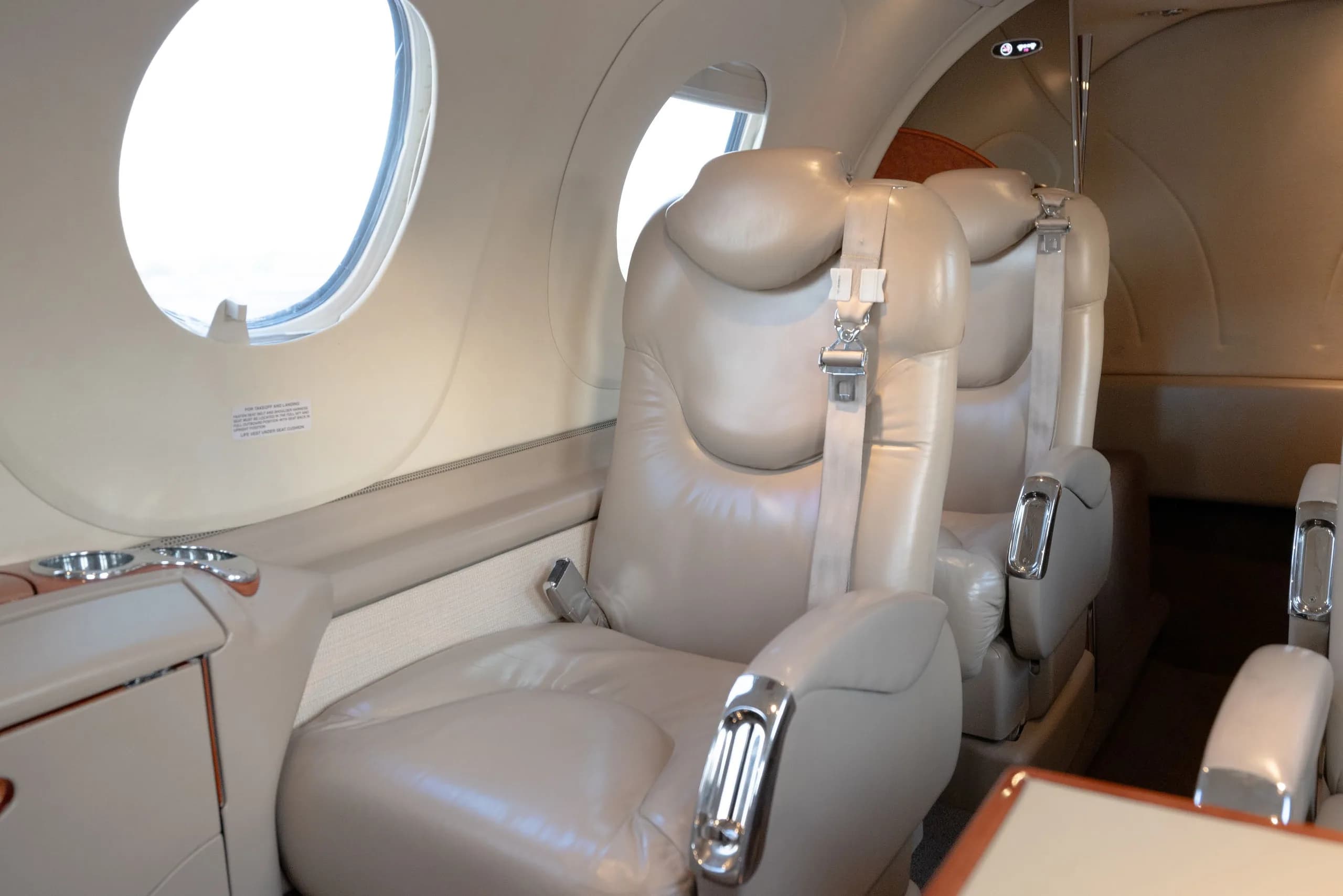 A close-up of light tan leather seats with seatbelts in the cabin of a business jet, next to a large oval window.