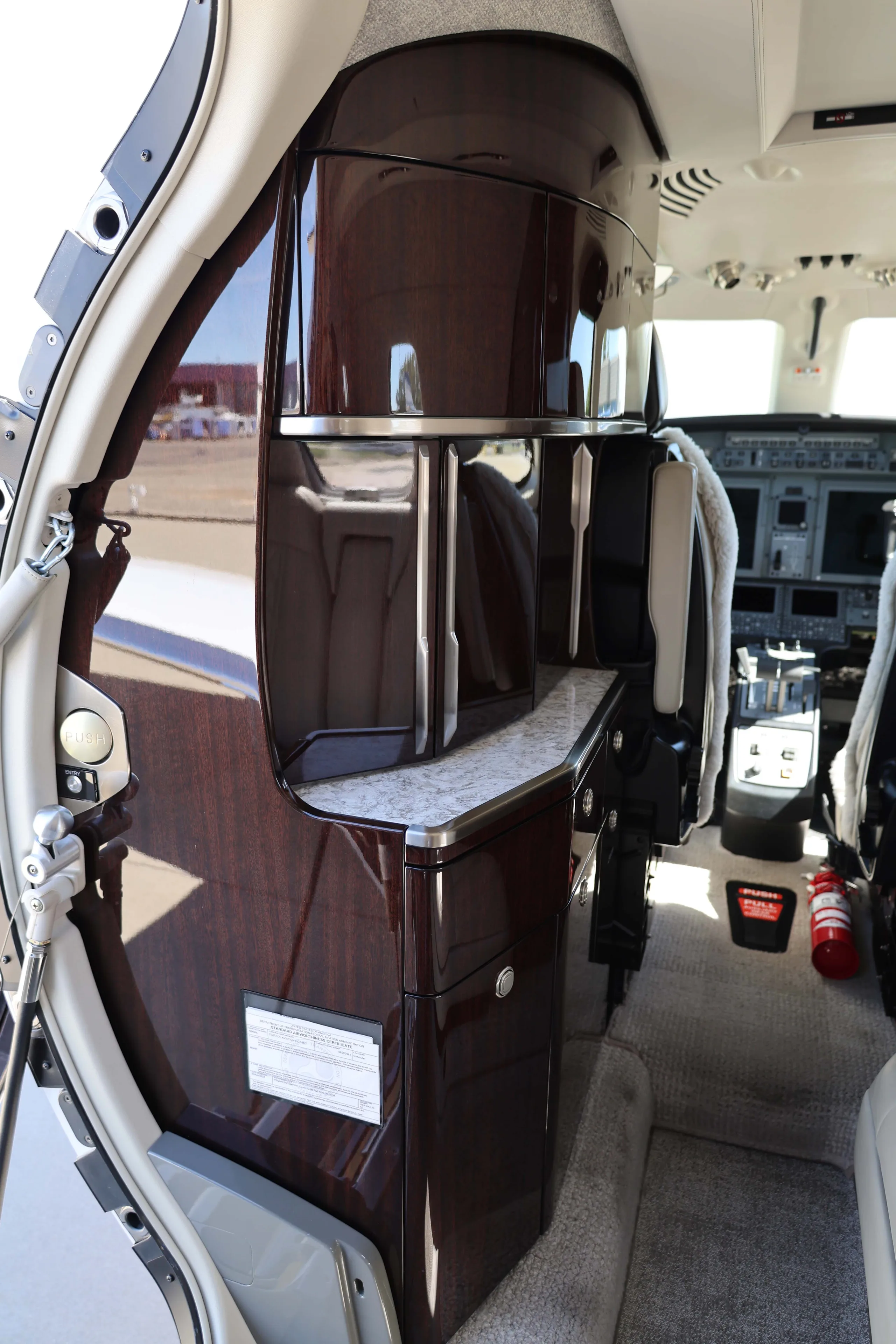 A close-up of the dark wood-paneled galley and storage area inside a private jet cabin, with the cockpit visible in the background.