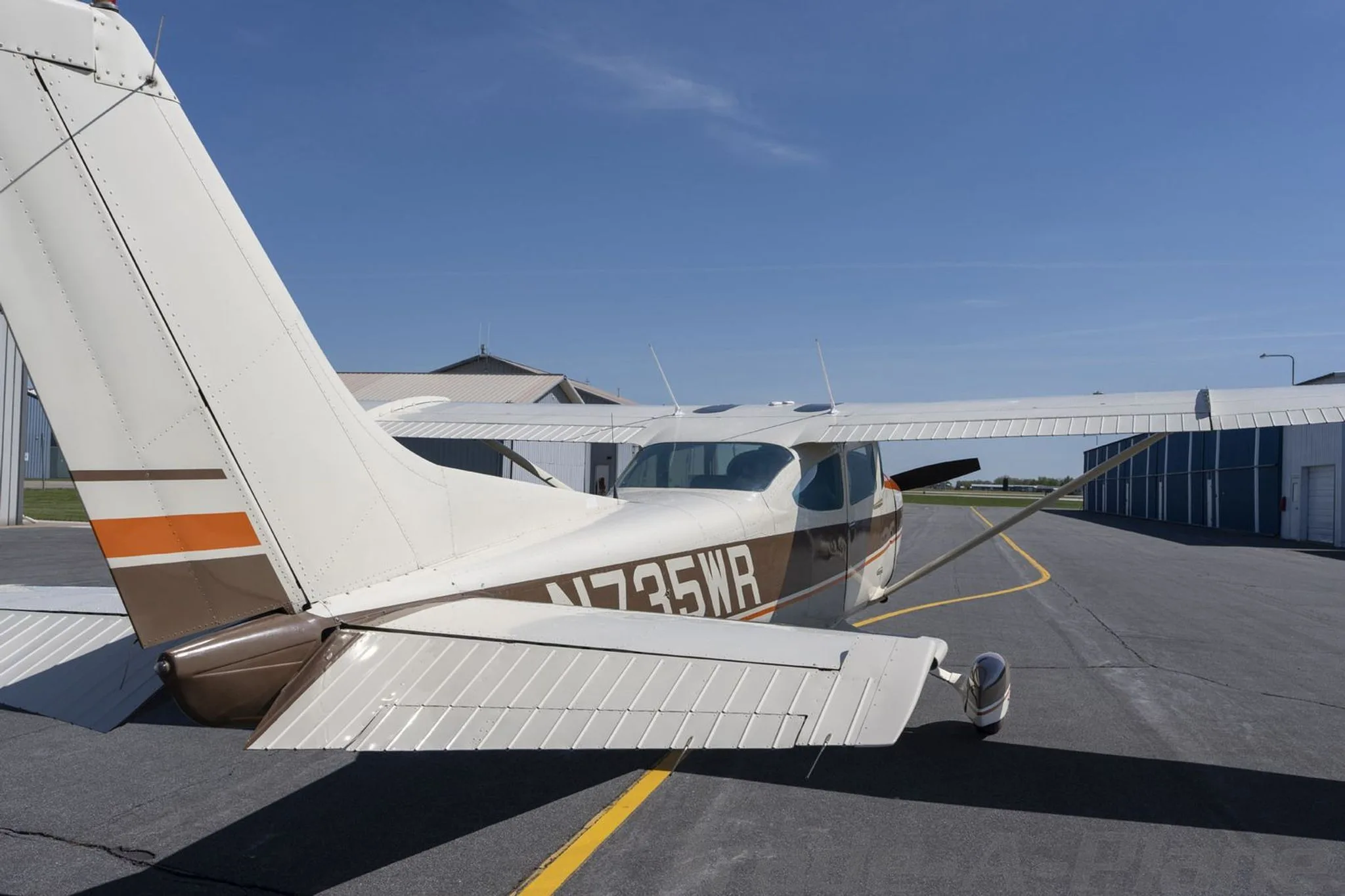 A rear three-quarter view of a white and brown Cessna 172 showing the tail and right wing, parked on an asphalt ramp.