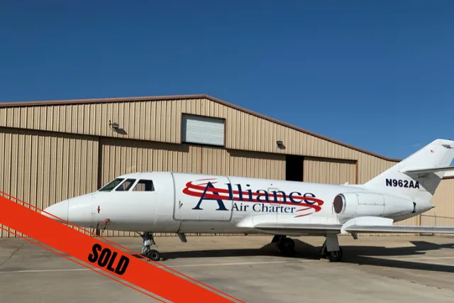 A white business jet with 'Alliance Air Charter' livery and registration N962AA, parked on a tarmac in front of a hangar under a clear blue sky.