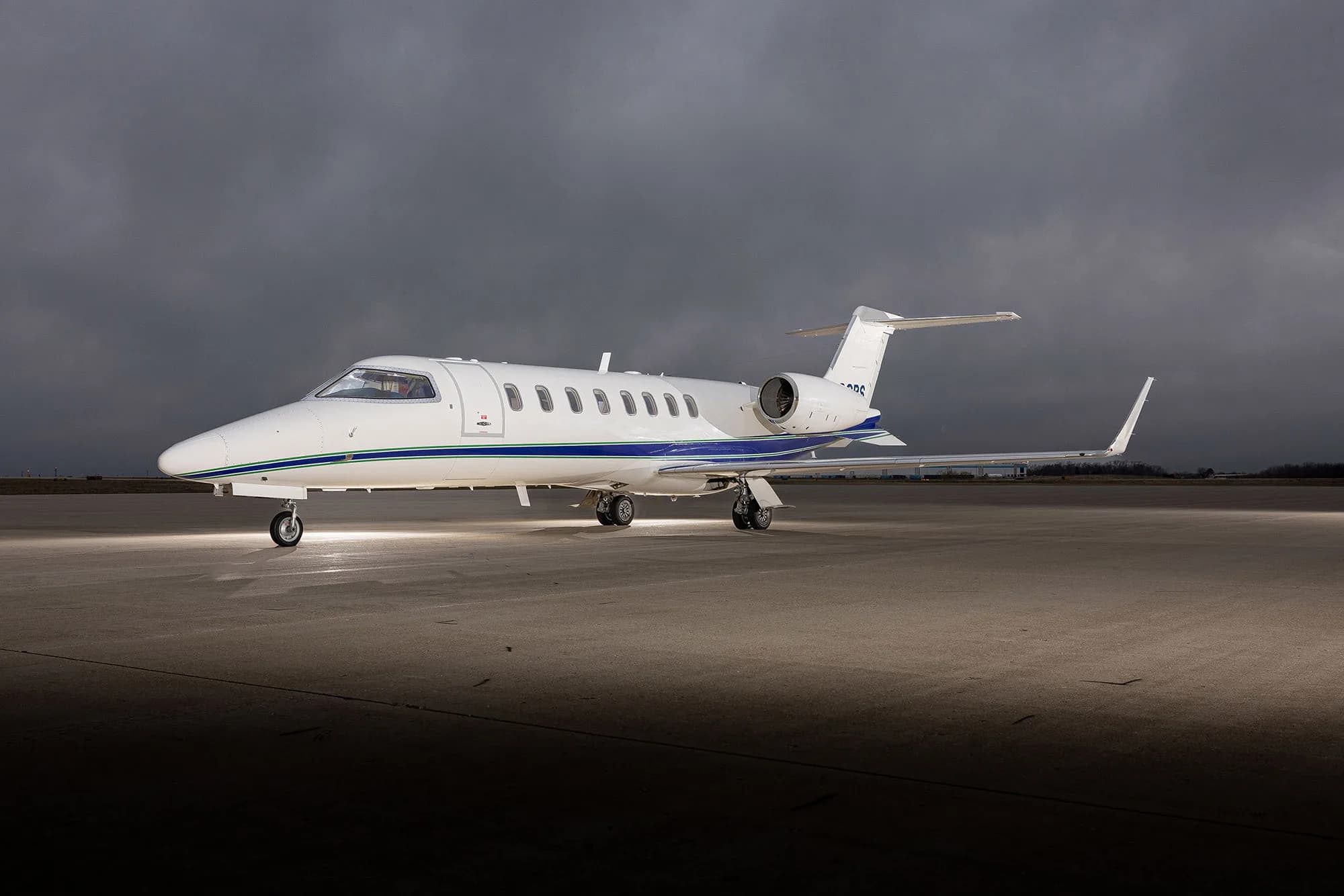 A white Learjet 75 with blue and green stripes parked on an tarmac under dramatic cloudy skies, viewed from the front-left.
