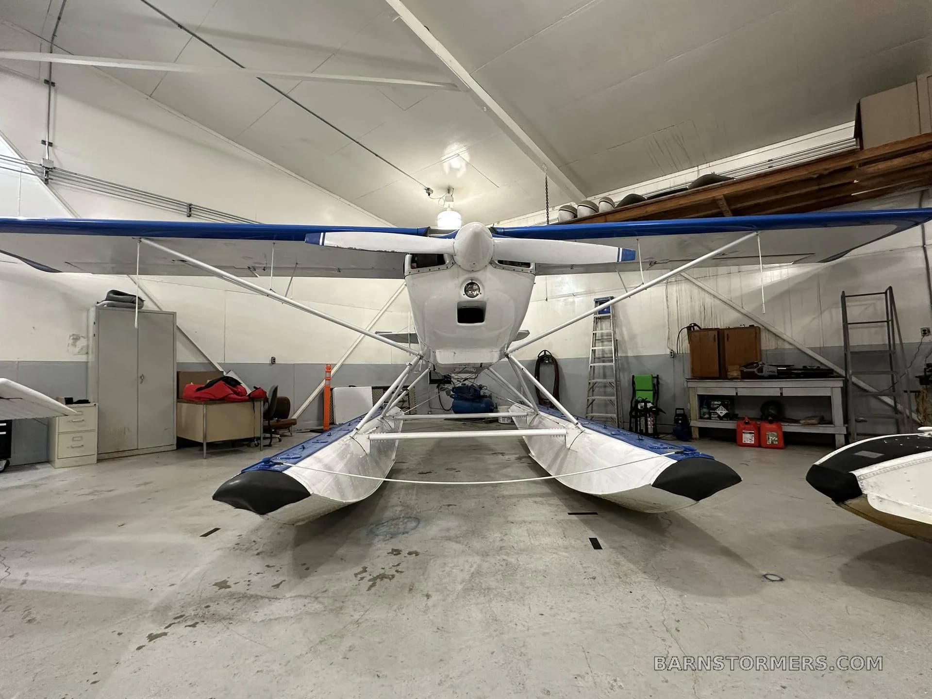 A blue and white Citabria floatplane is parked in a hangar, viewed from the front.