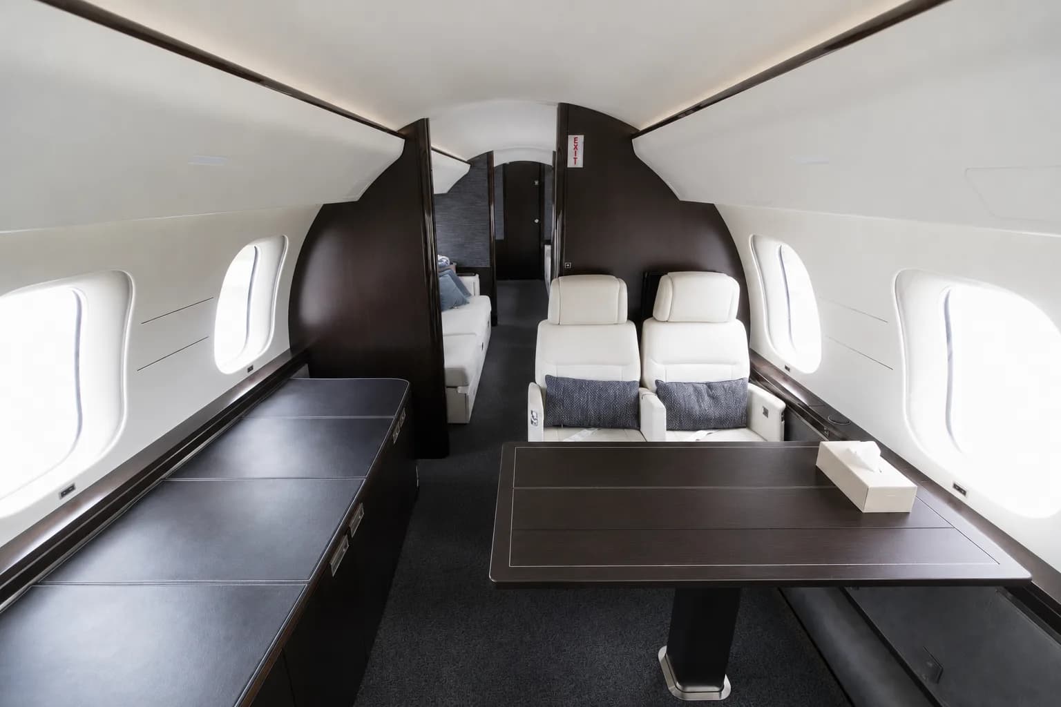 A section of a private jet cabin with a dark wood table, two white leather seats, and a view towards the rear.