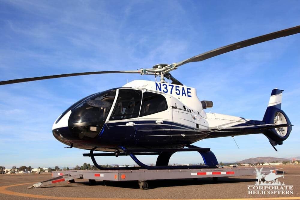 A white and dark blue helicopter with registration N375AE sits on a transport trailer on an airfield under a blue sky.