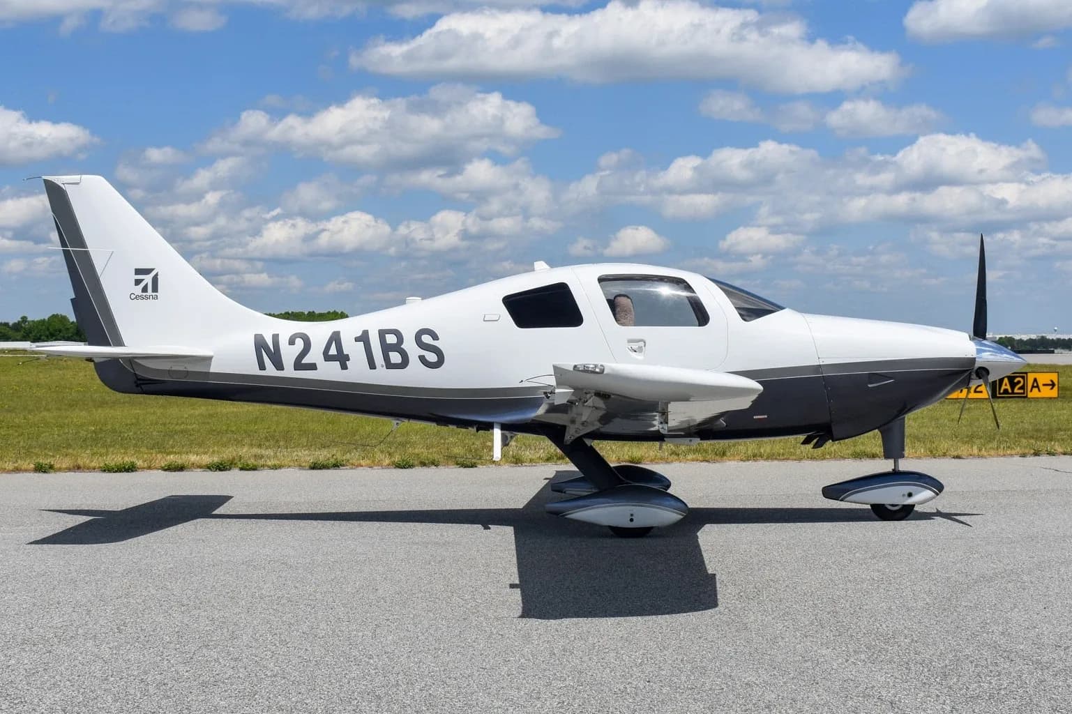A white and dark grey Cessna TTx (Corvalis 400) aircraft, N241BS, parked on an airfield under a partly cloudy sky.