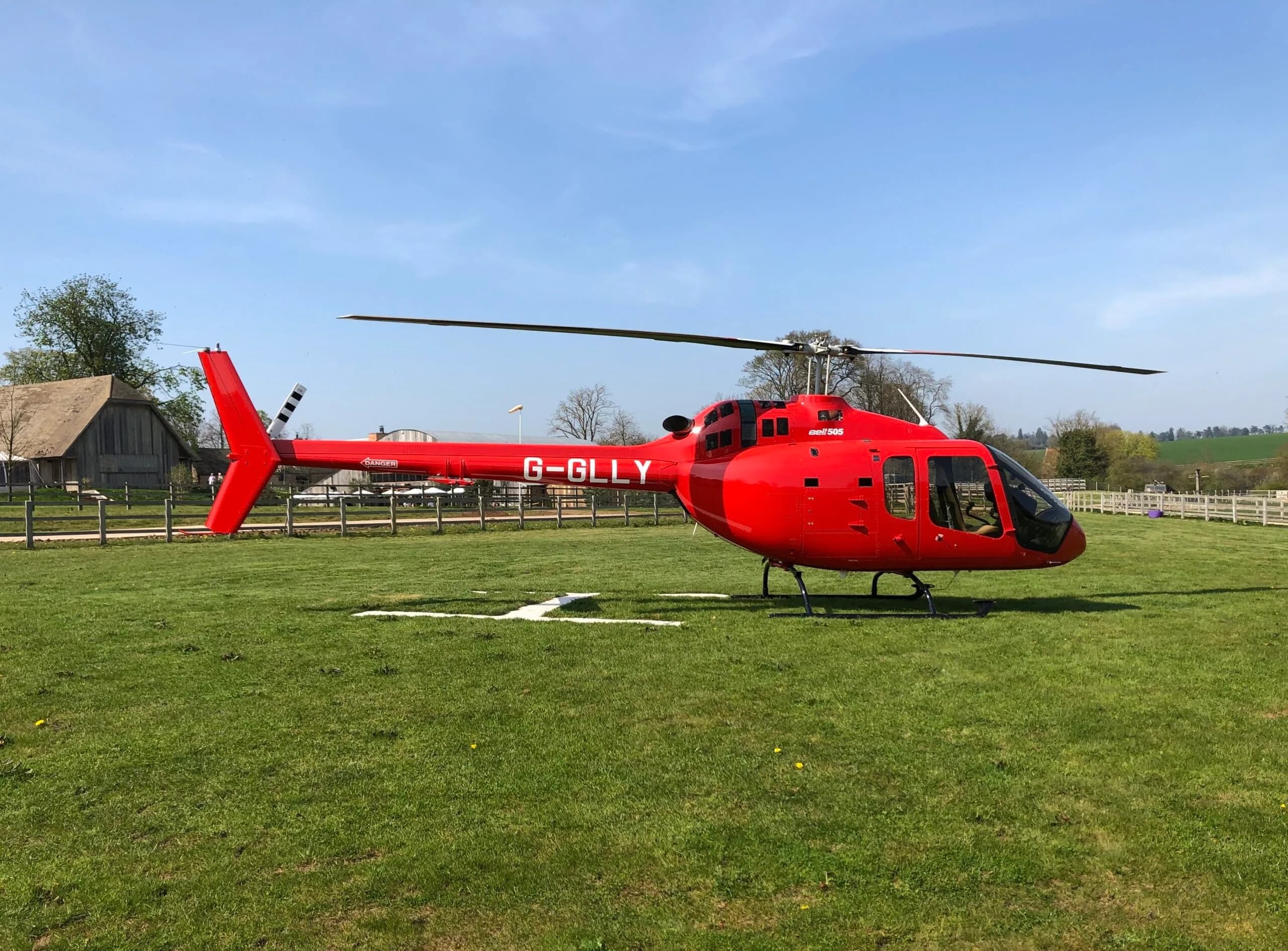 A red Bell 505 helicopter, registration G-GLLY, rests on a green field under a clear blue sky.