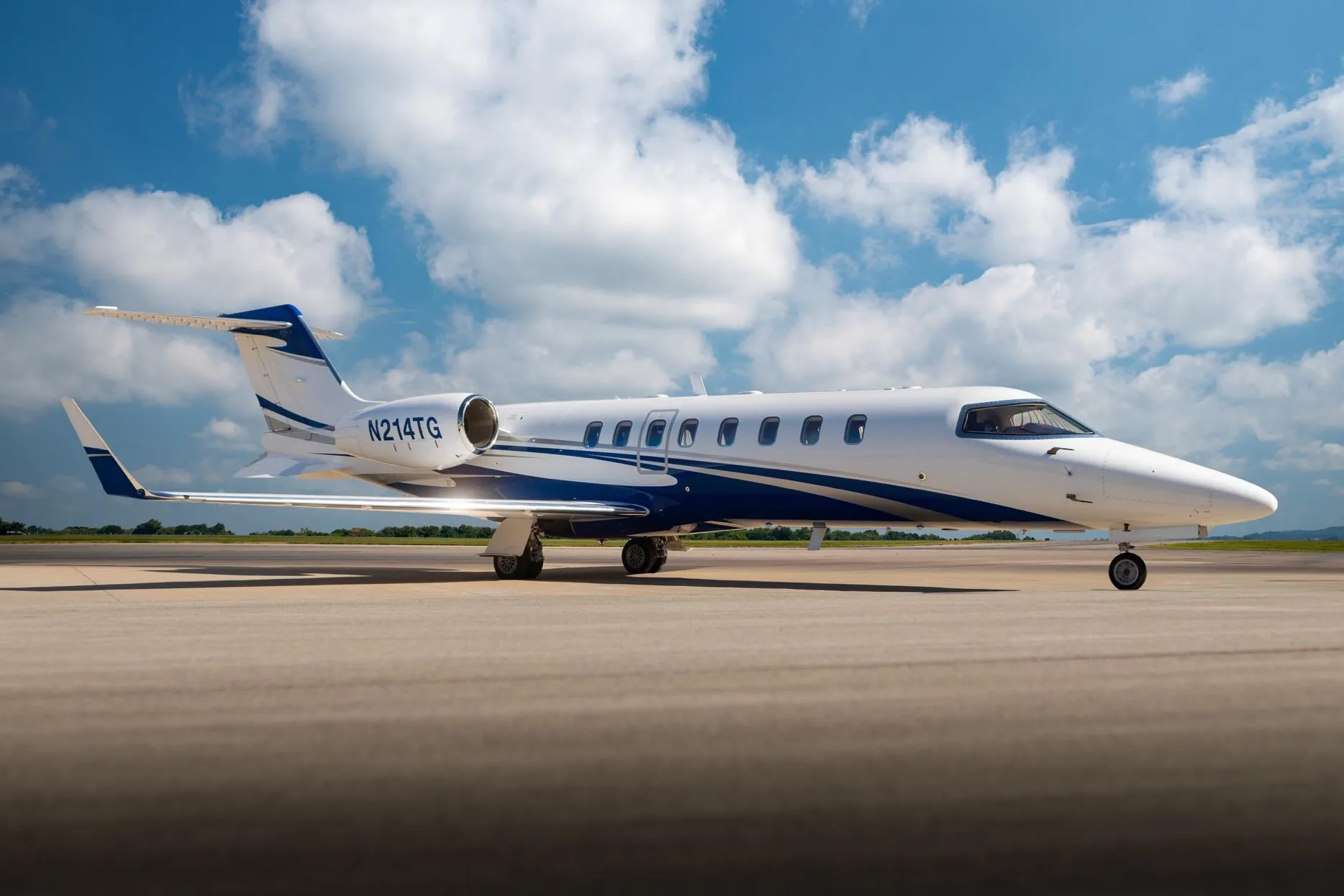 Side view of a white and blue Learjet 75 business jet, registration N214TG, on a tarmac under a blue sky.