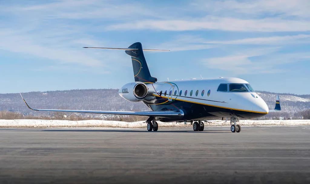 A Gulfstream G550 aircraft, registration N38GL, with a white and dark blue paint scheme, is parked on an airfield with snow-covered hills in the background.