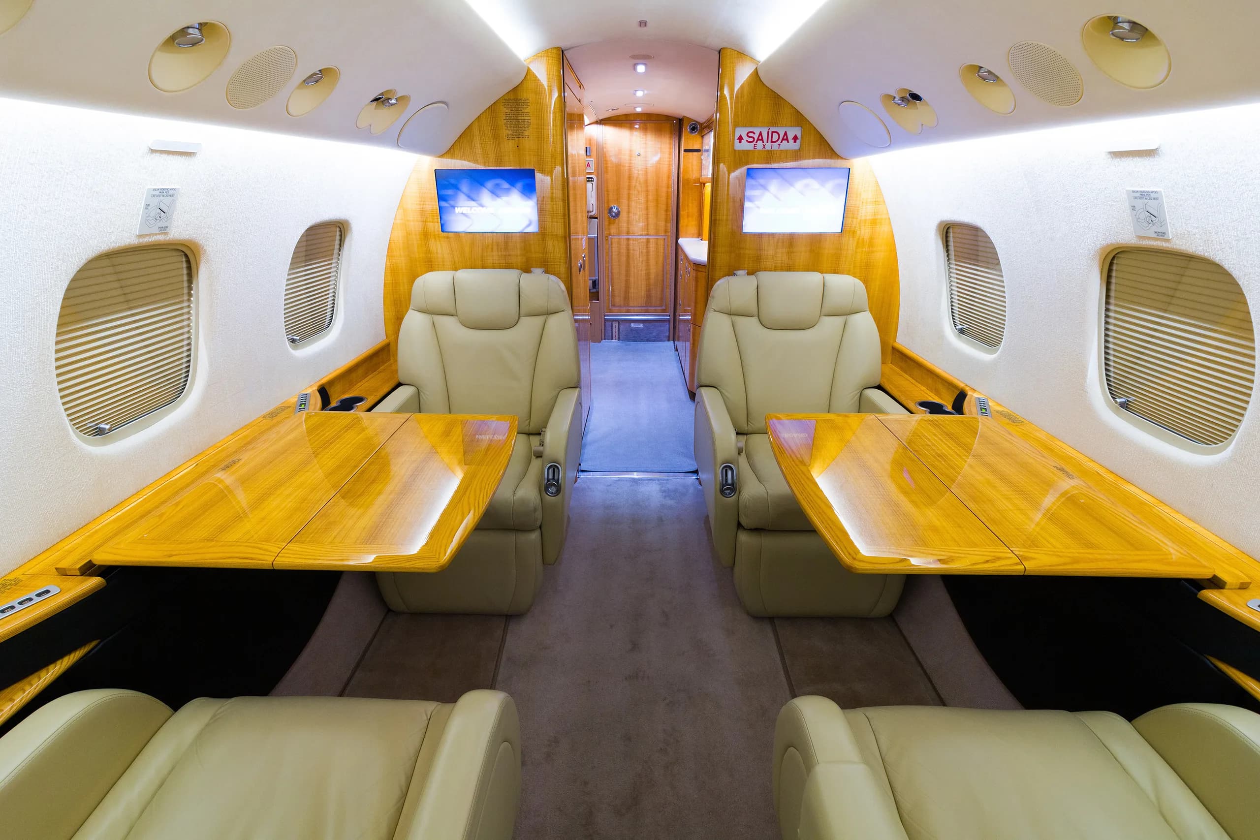The luxurious interior of a private jet cabin with two sets of facing beige leather seats and polished wooden tables.