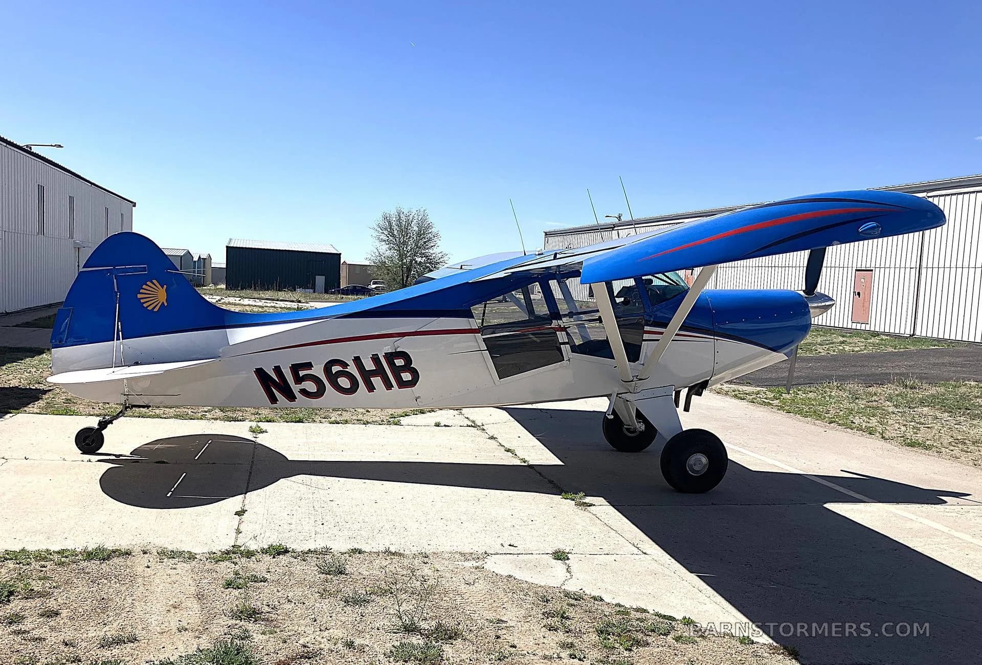 A blue and white high-wing aircraft with red stripes and tail logo, registration N56HB, parked on an asphalt apron under a clear blue sky.