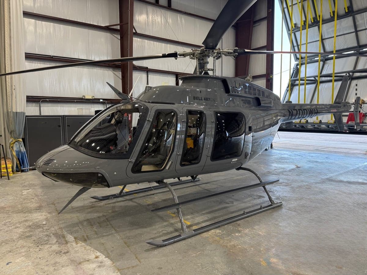 A grey Bell 407GXi helicopter is parked inside a hangar, viewed from the front-right side.