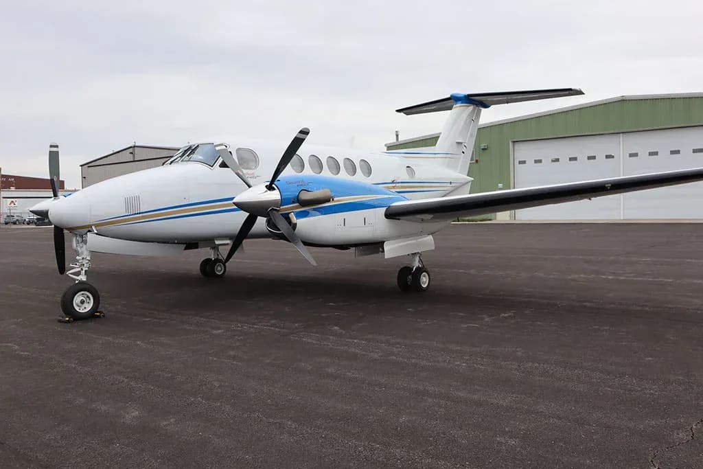 A white and blue twin-engine turboprop aircraft, likely a King Air, parked on an asphalt tarmac with hangars in the background.