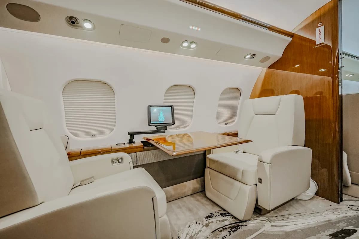 A section of a private jet cabin with two light-colored seats, a wooden table, and a small monitor.