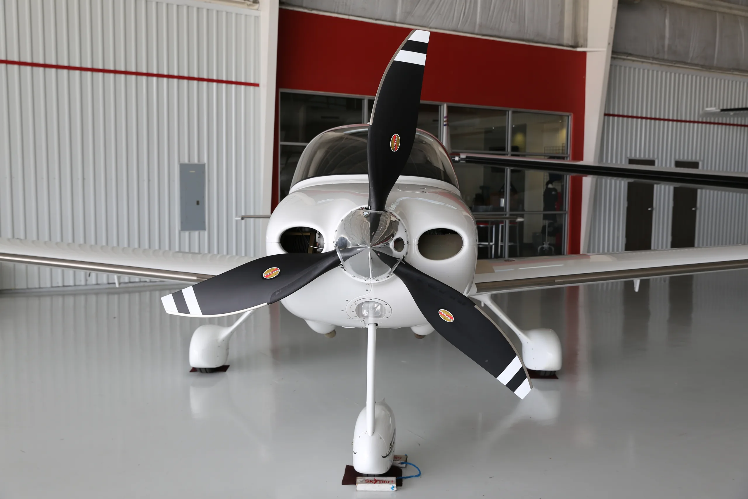 A head-on view of the white Cirrus SR22T aircraft with its three-bladed propeller, parked in a hangar.