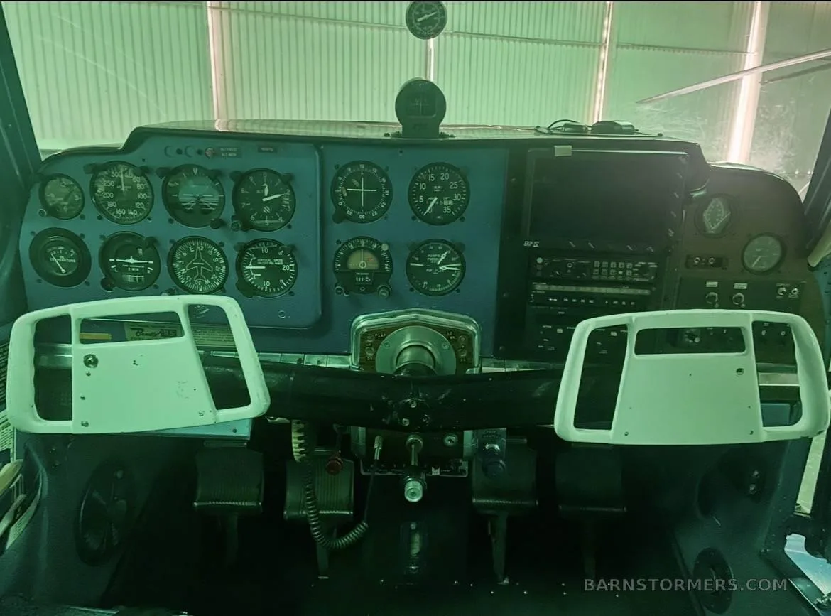 The blue instrument panel of a small aircraft with gauges and two white control yokes, viewed from inside a hangar.