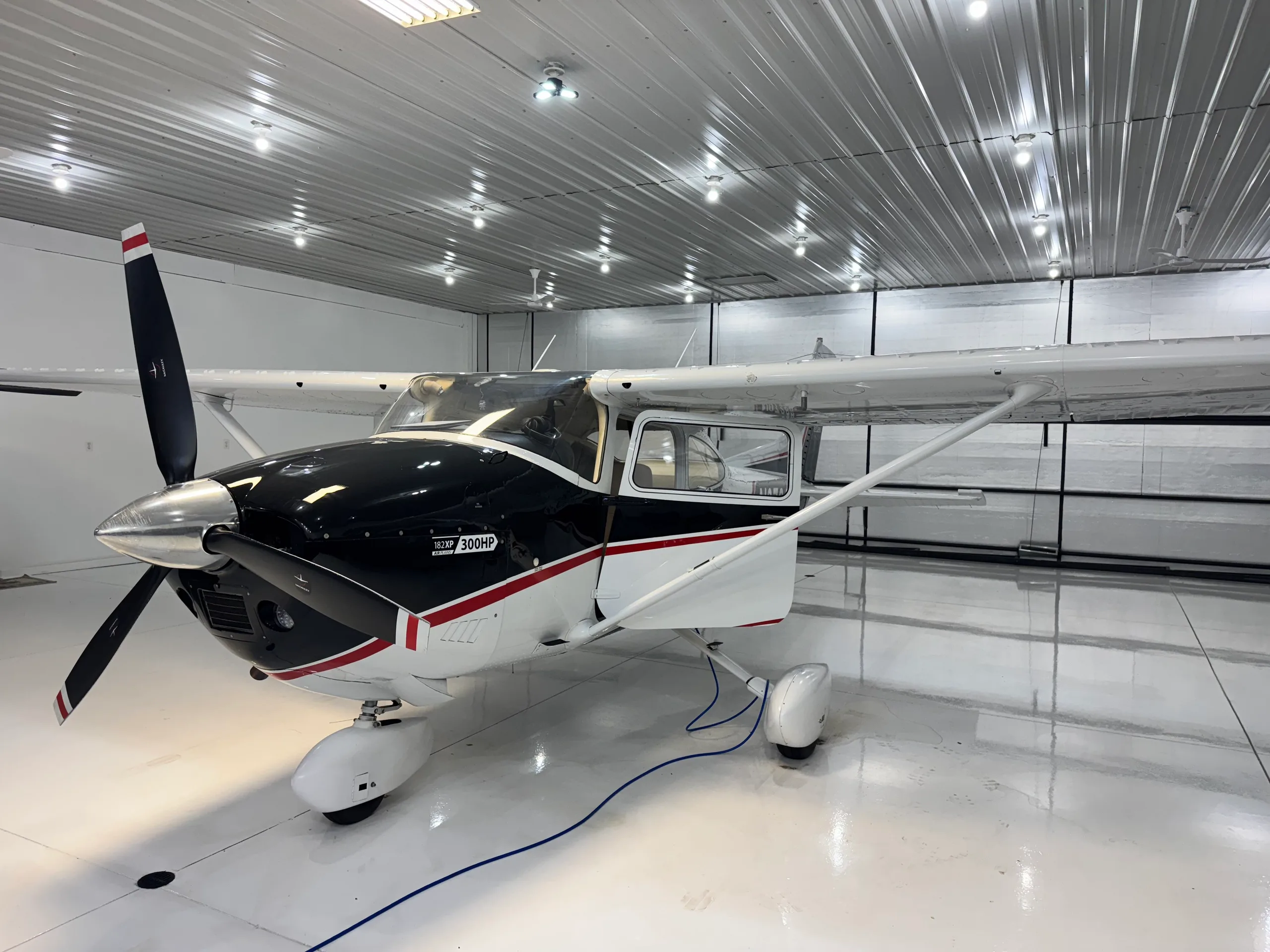A full front-right view of a black and white Cessna 182XP 300HP aircraft with a three-bladed propeller in a brightly lit hangar.