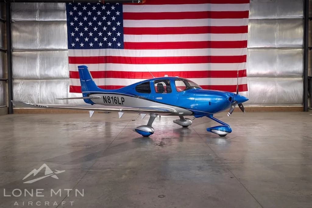 A blue and silver Cirrus SR22 aircraft, registration N816LP, parked in a hangar with an American flag in the background.