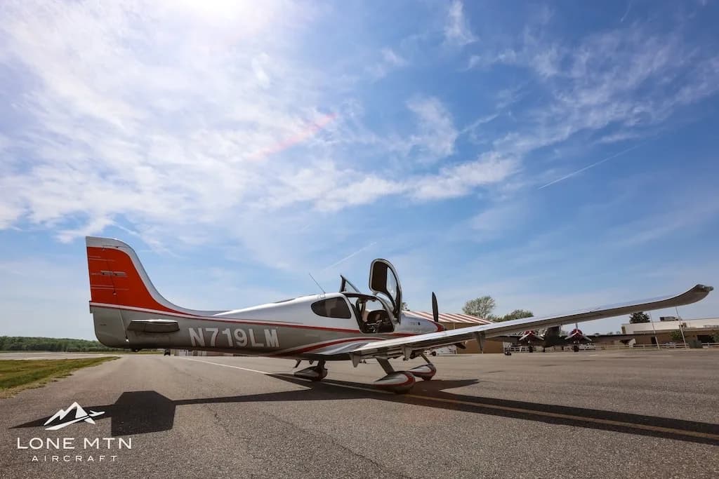 Low angle side view of a silver and orange Cirrus SR22 aircraft with doors open, parked on an asphalt runway under a blue sky.