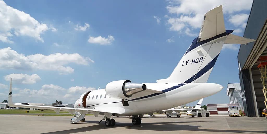 A white business jet with dark blue and grey stripes, registration LV-HQR, is parked on a tarmac with its passenger door open and stairs extended, next to a hangar.