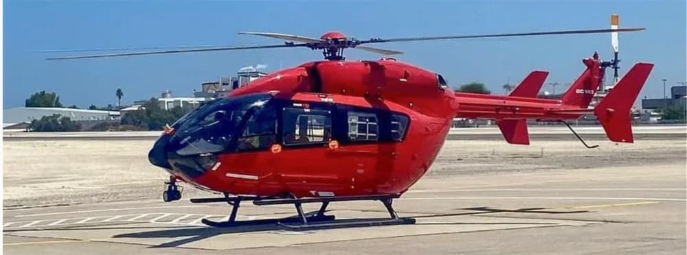 A red Airbus H145 helicopter is parked on a tarmac with markings under a clear sky.