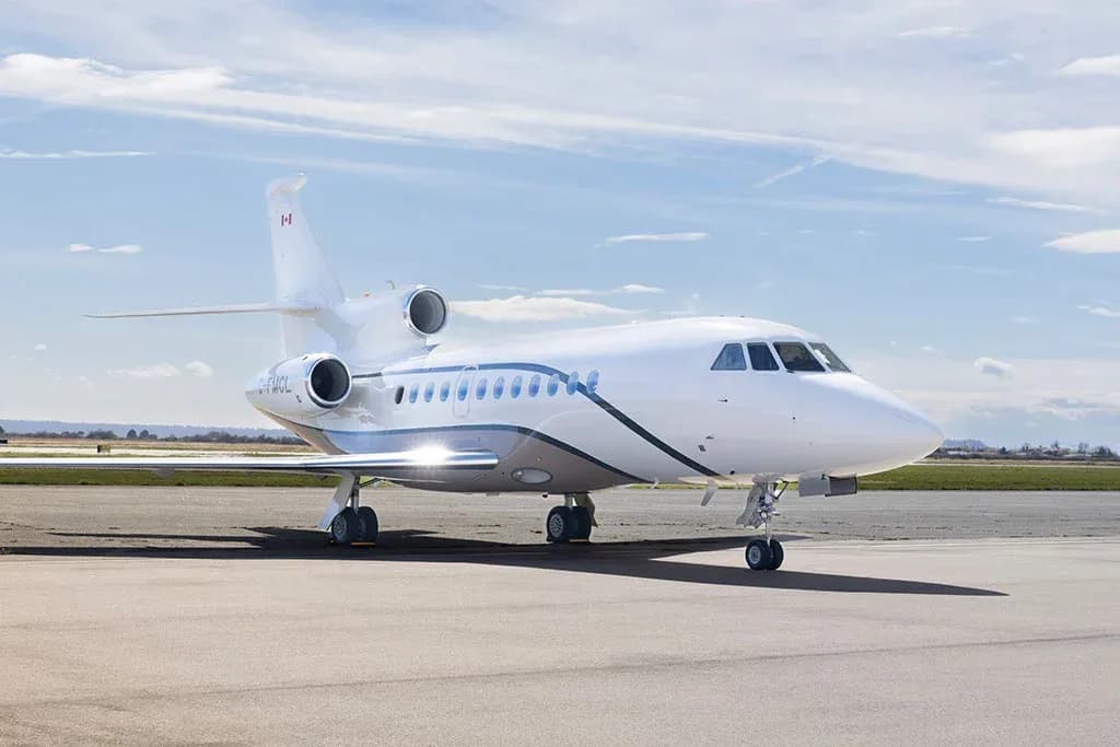 A white Dassault Falcon business jet with a brown stripe, registration C-FMCL, parked on an airfield under a partly cloudy sky.