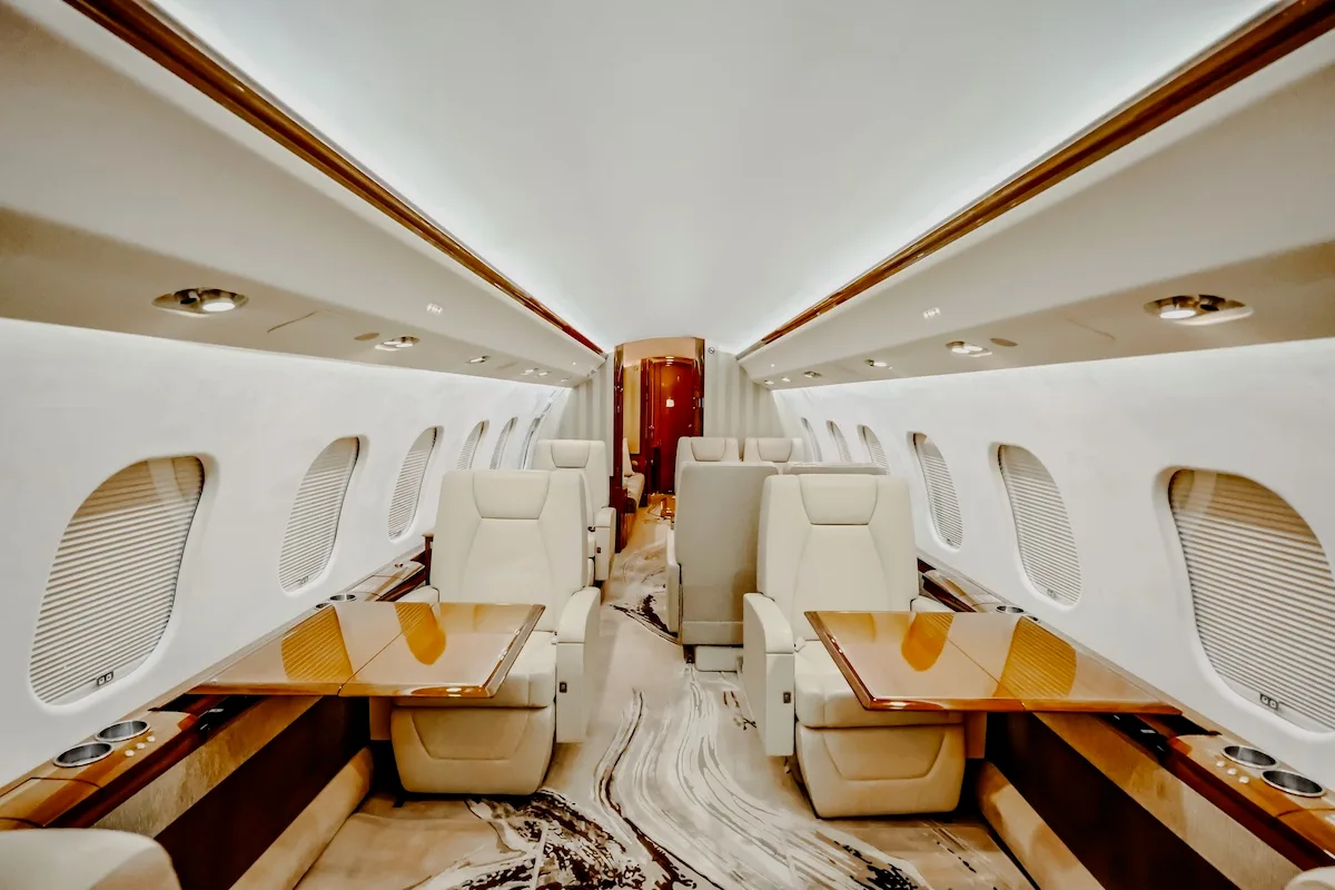 A view down the aisle of a private jet cabin with light-colored seats and wooden tables.