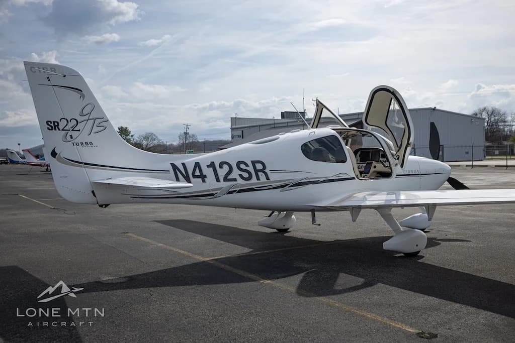 A white Cirrus SR22T aircraft with open doors is parked on an asphalt runway next to airport buildings.