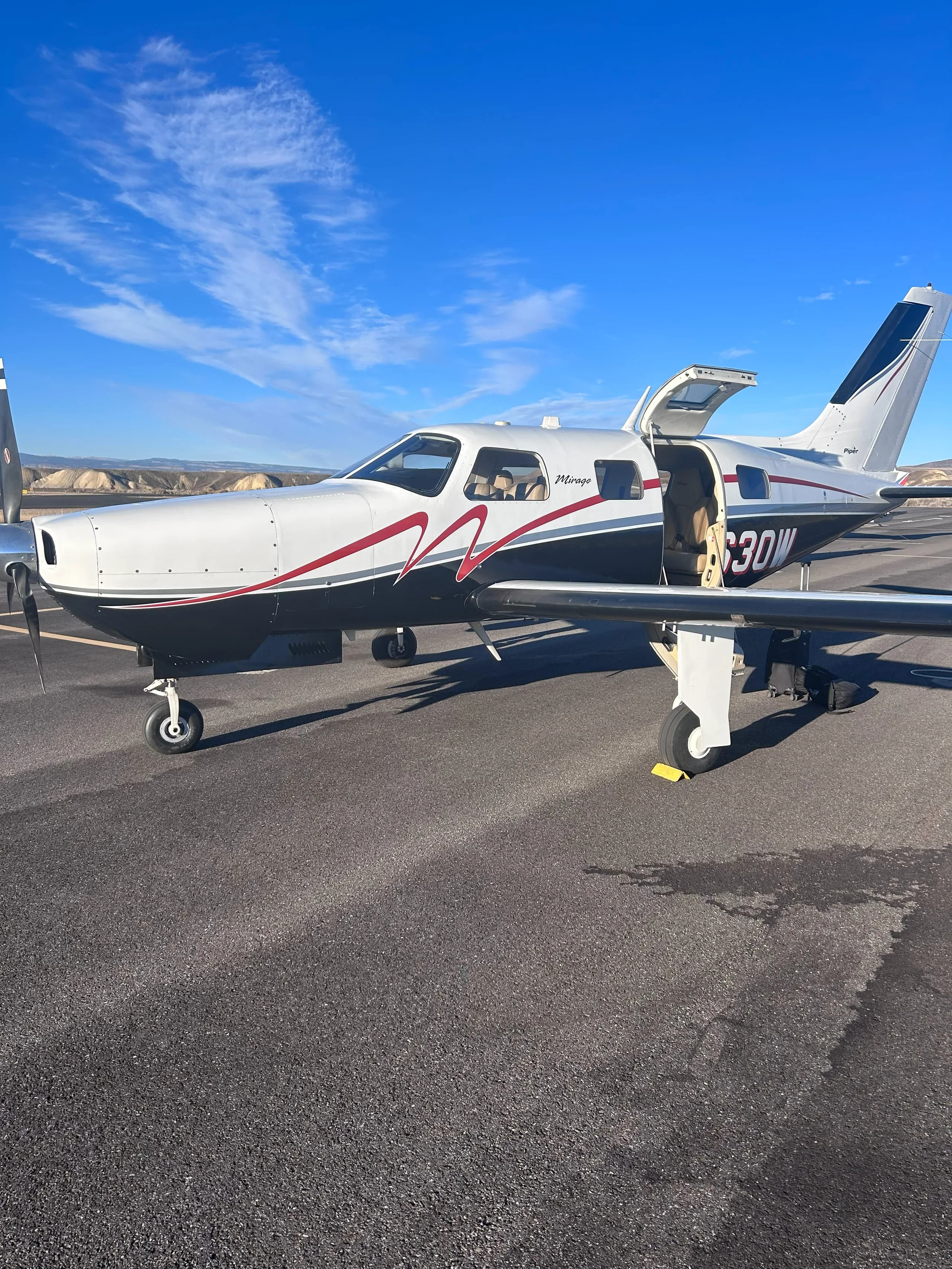 The Piper Mirage N1630W, white and black with red trim, parked on an airfield with its door open under a clear blue sky.