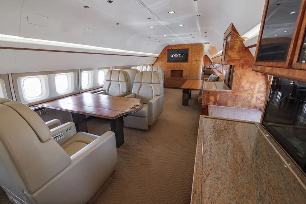 The luxurious interior cabin of an aircraft, featuring beige seats, wooden tables, and a screen displaying the Avjet logo.