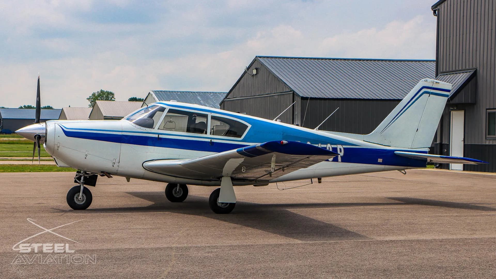 A side view of a white and blue single-engine aircraft parked on a tarmac with a hangar behind it.