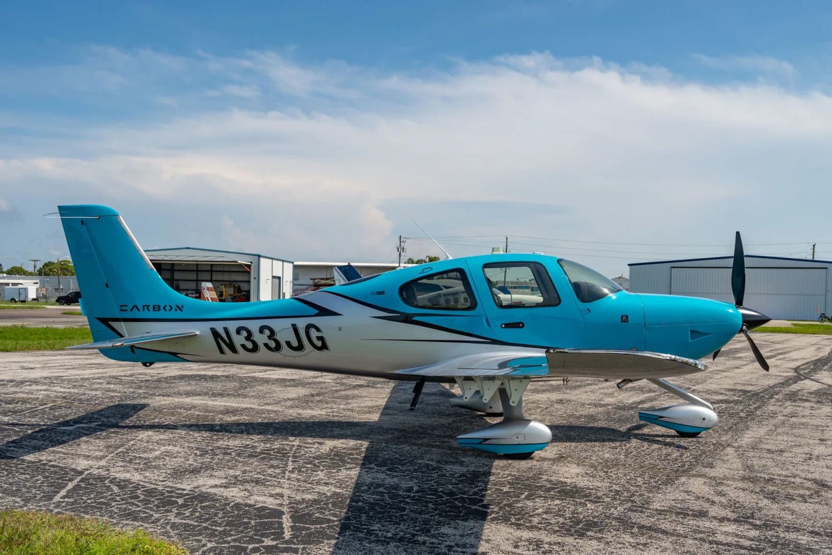 A blue and silver Carbon aircraft (N33JG) parked on an asphalt ramp with hangars in the background.